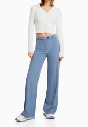 Biały krótki kardigan z zapięciem na guziki, w połączeniu z miękkimi niebieskimi spodniami z szerokimi nogawkami, które mają zapięcie na guziki z przodu i gładką teksturę. Szare trampki.