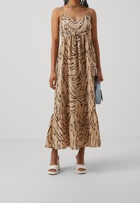 Vestido maxi en beige con rayas abstractas negras, con tirantes finos y cintura fruncida, que se ensancha en el dobladillo. Combinado con zapatos de tacón blanco.