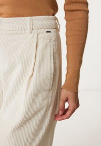 Cream corduroy trousers with vertical ribbed texture, elastic waistband, and side pockets, paired with a fitted brown long-sleeve top.