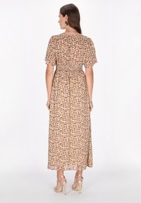 Maxi jurk in lichtbeige met bloemenprint, korte mouwen, smock taille en vloeiende rok. Draagt strappy beige hakken.