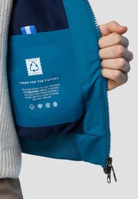 Interno di una giacca isolante blu con una tasca, caratterizzato da un logo di riciclaggio e un testo sui fili riciclati, tessuto testurizzato e chiusura con zip.