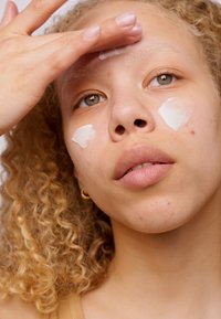 Gros plan d'une personne appliquant une crème blanche sur son visage, avec des cheveux bouclés, une peau claire et des imperfections visibles. Boucles d'oreilles en or en forme de cerceau visibles.