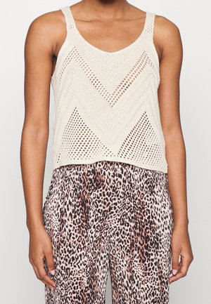 Frau trägt ein ärmelloses beiges Stricktop mit Chevron-Mustern und hoch taillierte, plissierte Hosen mit Leopardenmuster.