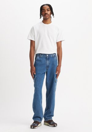 White crew-neck t-shirt paired with loose-fitting blue cargo jeans featuring side pockets. Footwear includes brown and patterned sneakers.