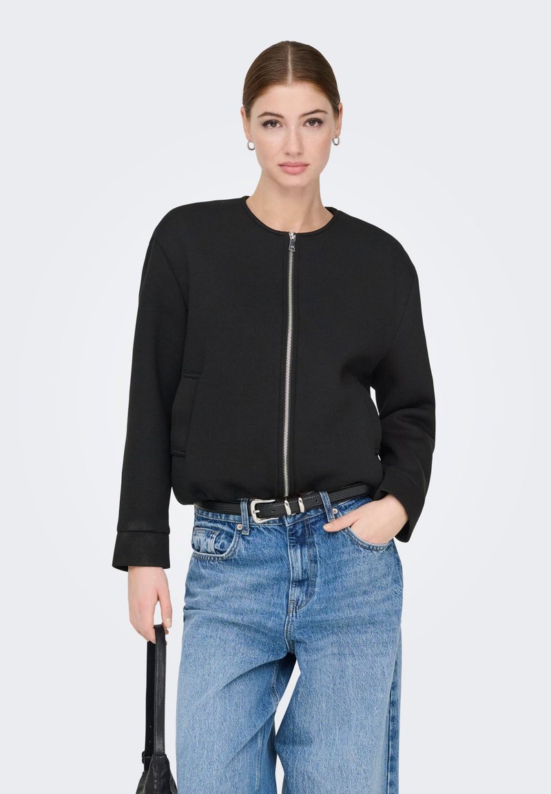 Femme aux cheveux tirés en arrière portant une veste noire à fermeture éclair et un jean bleu, tenant un sac à main noir dans une main, l'autre main dans la poche, fond neutre.