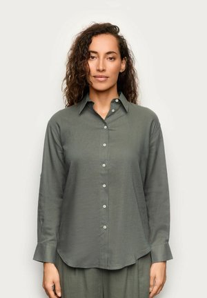 Donna con capelli ricci che indossa una camicia grigia a maniche lunghe con bottoni e pantaloni abbinati, in piedi davanti a uno sfondo chiaro e uniforme.