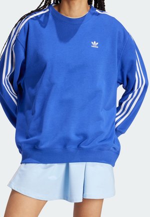 Osoba nosząca niebieską bluzę Adidas z białymi paskami na rękawach oraz jasnoniebieską plisowaną spódnicę.