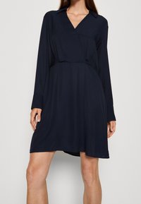 Navyblaues Kleid mit V-Ausschnitt, langen Ärmeln und lockerer Passform, aus einem glatten Stoff mit einem knielangen Saum.