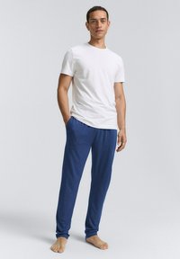 T-shirt blanc à col rond et pantalon de détente bleu. Les pantalons ont une taille élastique et une coupe ample. Le modèle se tient pieds nus sur un fond clair.