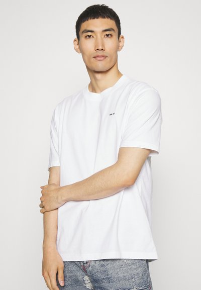 Weißes Baumwoll-T-Shirt mit kurzen Ärmeln und Rundhalsausschnitt, mit einem kleinen schwarzen Logo auf der linken Brust. Das Modell trägt eine helle Denim-Jeans.
