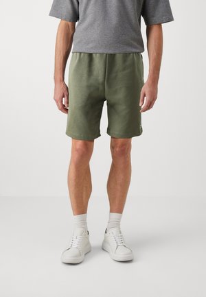 Mann trägt olivgrüne Shorts, graues T-Shirt, weiße Crew-Socken und weiße Sneakers und steht vor einem schlichten hellen Hintergrund.