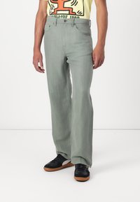 Green wide-leg jeans made of denim, featuring a high waist and five-pocket design, paired with black sneakers and a yellow graphic t-shirt.