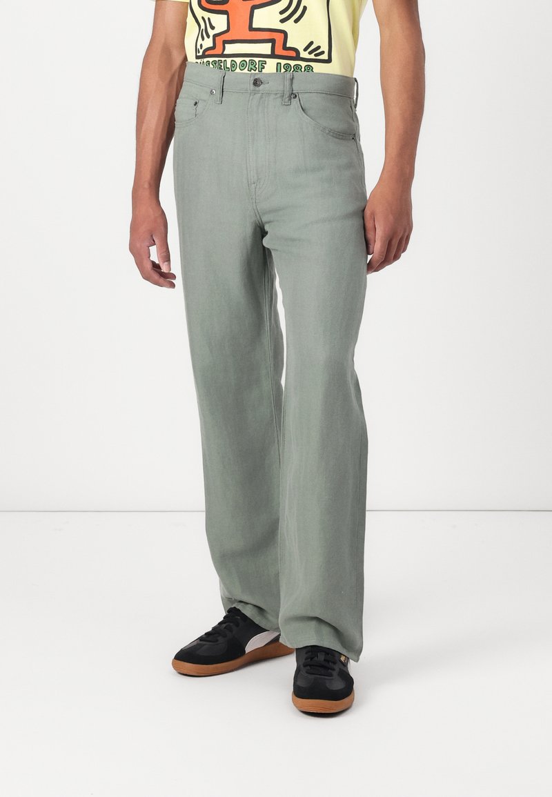 Green wide-leg jeans made of denim, featuring a high waist and five-pocket design, paired with black sneakers and a yellow graphic t-shirt.