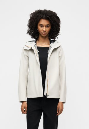 Jeune femme aux cheveux bouclés portant une veste gris clair zippée sur un haut noir et un pantalon noir, debout devant un fond uni.