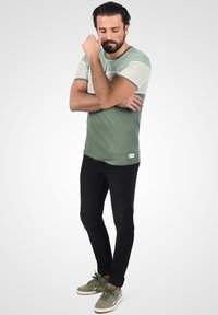 Grøn og grå kortærmet skjorte med rund hals, parret med sorte slim-fit jeans og grågrønne sneakers. Har et logo patch.
