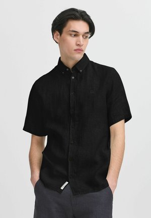 Černá krátkosleeve košile s knoflíky vyrobená z lehké tkaniny se subtilní texturou a malým logem na hrudi, spárovaná s tmavými kalhotami.