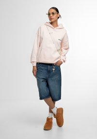 Rosa hettegenser med frontlomme, slitte denimshorts og brune ankelstøvler. Tilbehøret inkluderer klare solbriller. Enkel, moderne stil.