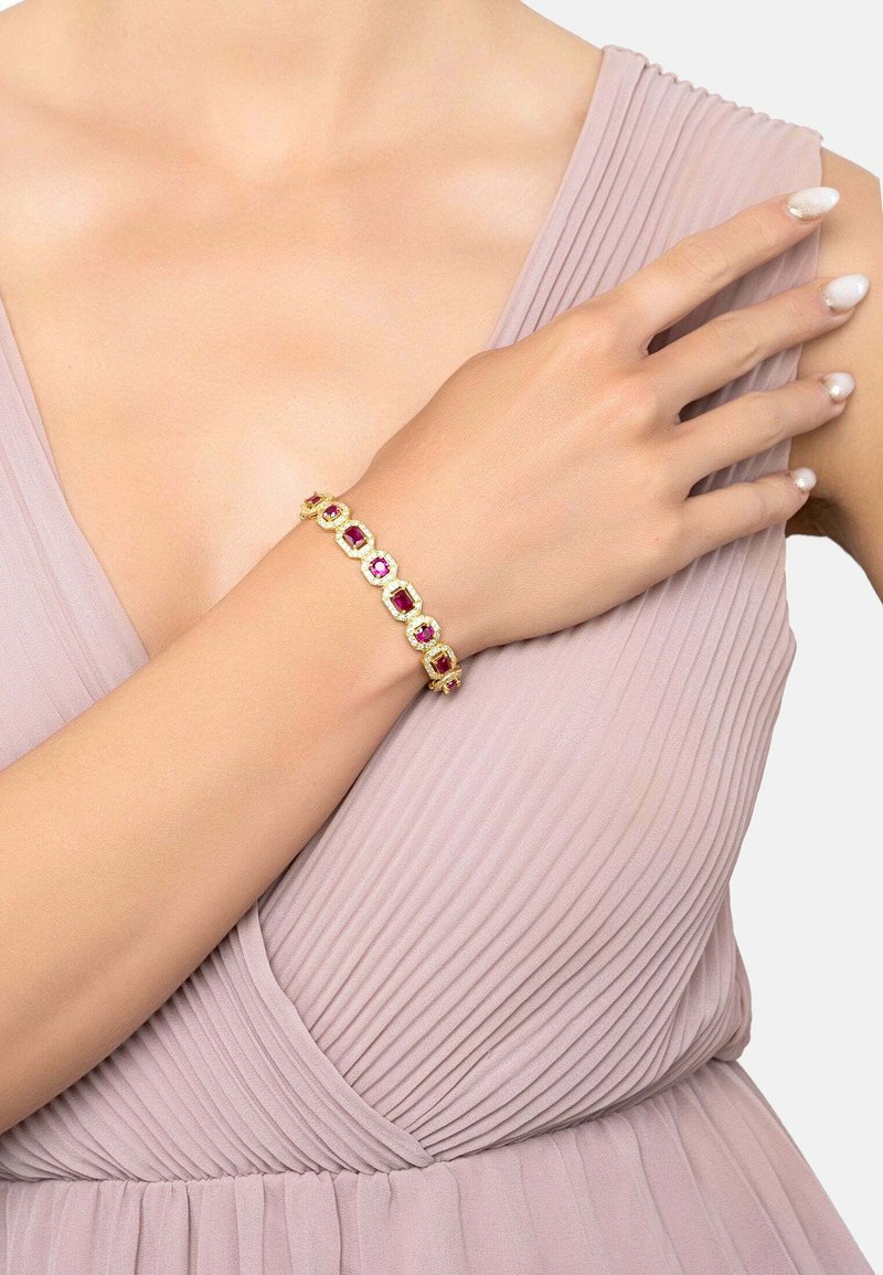 Gold bracelet with alternating pink and red gemstones, featuring a smooth, textured design, worn on a hand against a pleated dress background.