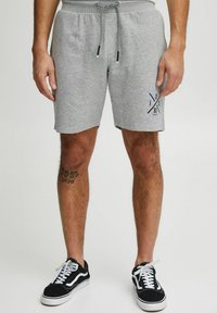 Lys grå bomulds shorts med elastisk talje og snortræk, med et mørkeblåt logodesign på venstre ben. Sorte sneakers med hvide accenter.