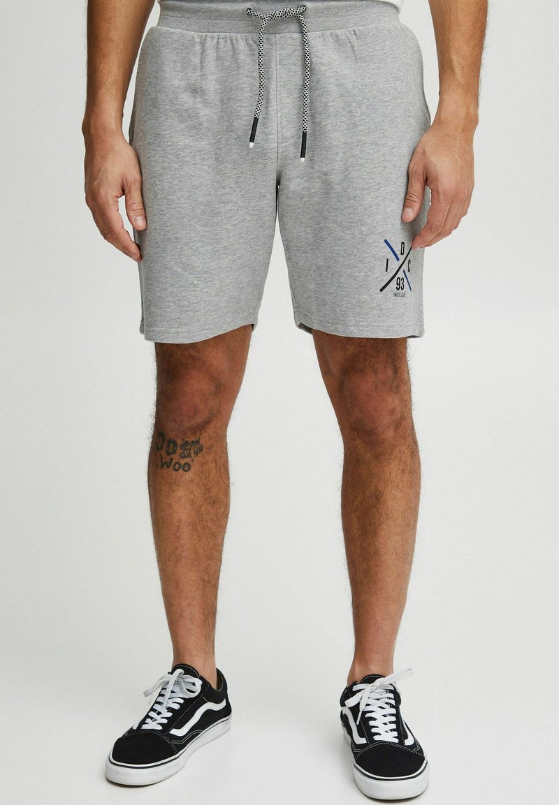 Lys grå bomulds shorts med elastisk talje og snortræk, med et mørkeblåt logodesign på venstre ben. Sorte sneakers med hvide accenter.