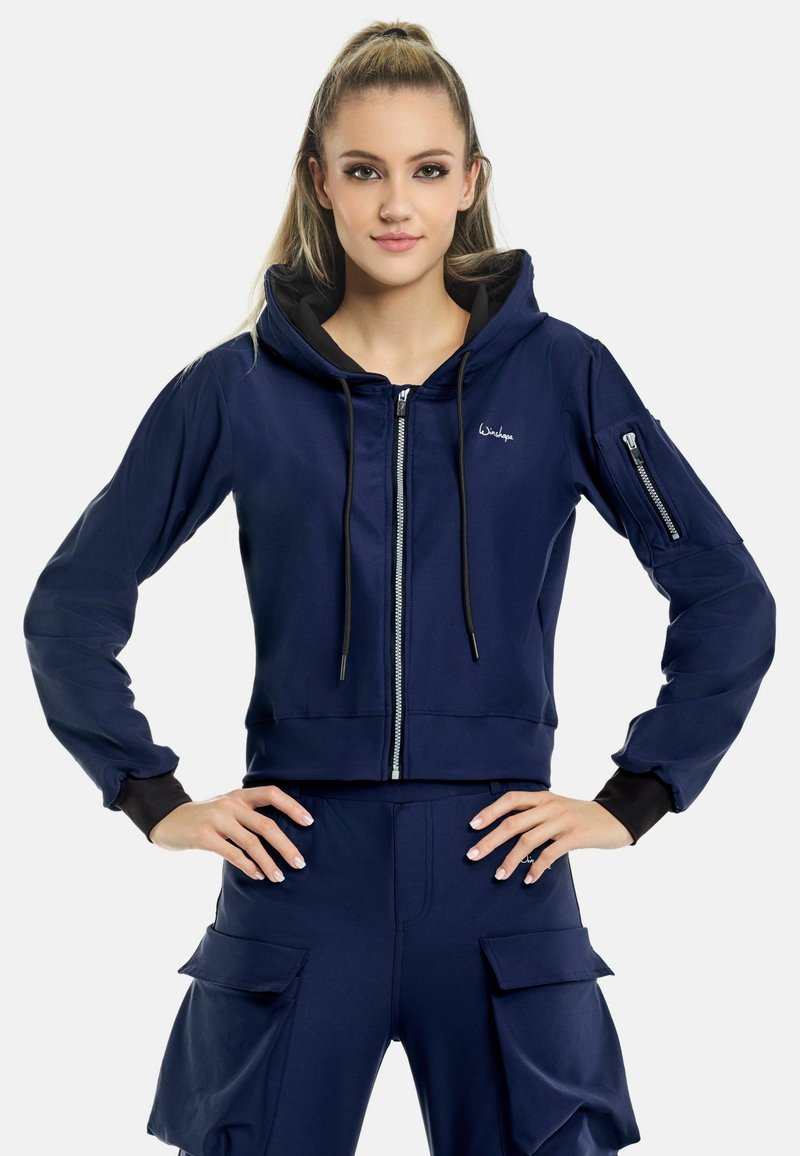 Marinblå kort hoodie med framdragkedja, justerbara snoddar och svarta detaljer. Har långa ärmar och en ficka med dragkedja på armen.