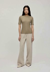 Beige tettskjorte med korte ermer, kombinert med lysebeige vidde bukser, begge i et glatt stoff. Modellen står foran en nøytral bakgrunn.