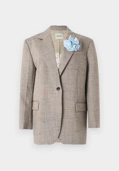 Blazer beige texturé à bouton unique avec col cranté, poches à rabat, et fleur en tissu bleu clair sur le revers gauche.