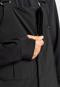 Hand die rits van een zwart uitrustingsbretel optrekt over een donker lange mouwen shirt met duimopening in de manchet.