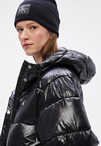 Melna pūkaina jaka ar spīdīgu apdari, ar kapuci un regulējamu auklu; aprīkota ar sudraba ražojuma detaļām un mazu logo uz krūtīm. Valkāta ar tumšu cepuri (beanie).