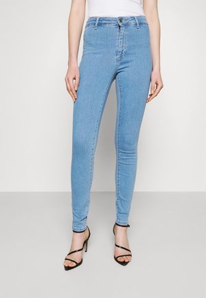 Jeans skinnies azul claro feitos de denim, com uma cintura média, fecho de botão e uma textura suave, combinados com sapatos de salto pretos com tiras.