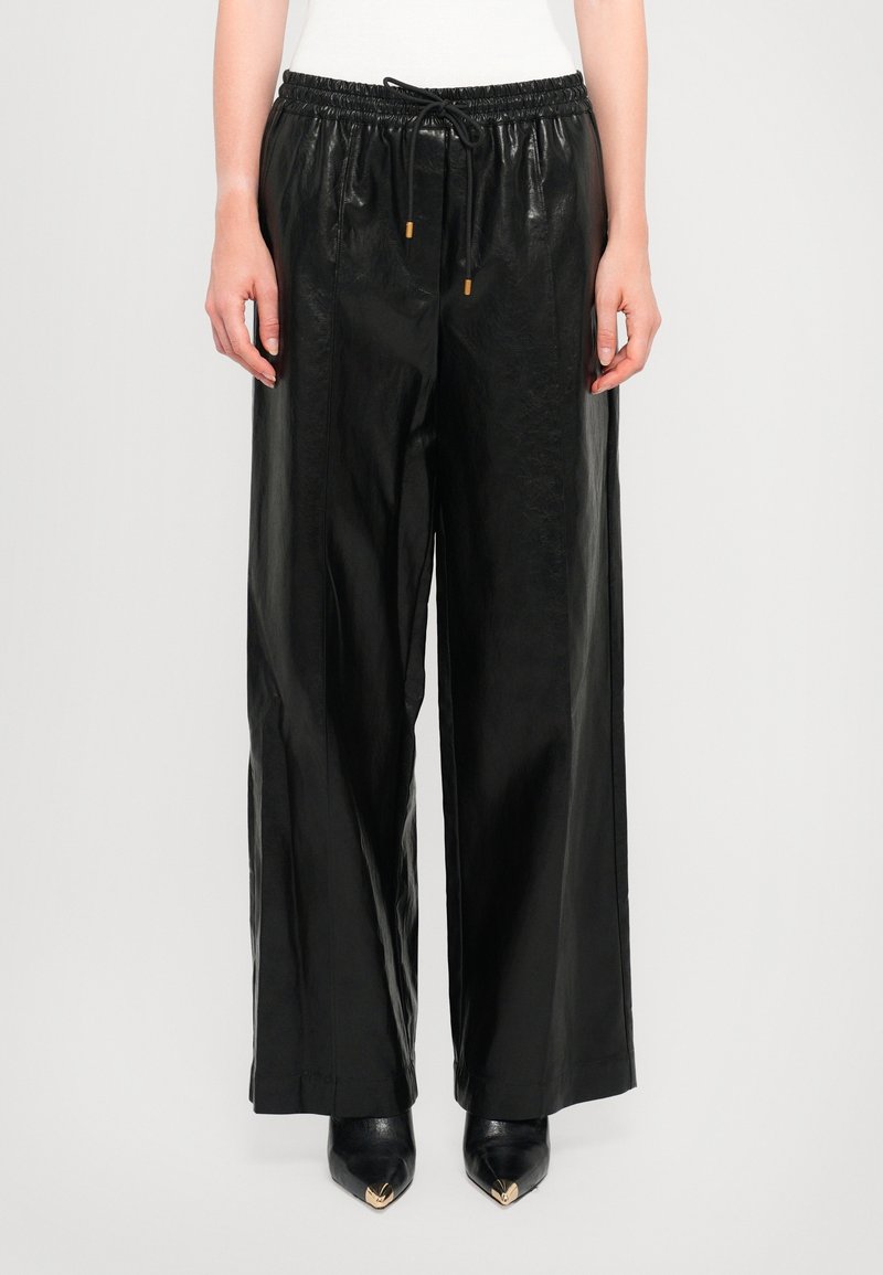 Pantalon large noir en similicuir avec taille élastique et cordon, porté avec des chaussures noires à bouts pointus et un haut blanc.