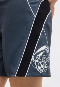 Σκούρο γκρι σορτς με ασπρόμαυρες ριγέ πλευρικές πινελιές και άσπρο γραφικό με κύμα και σερφίστας στο πλάι κοντά στο τελείωμα.