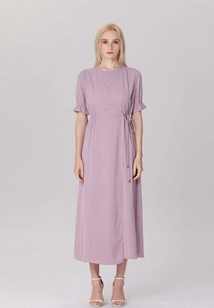 Femme blonde debout face à nous portant une robe portefeuille violet clair avec des manches courtes à volants et des talons blancs à bride cheville sur un fond uni.