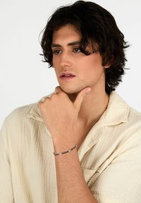 Bracciale in metallo con perline nere alternate a dettagli in argento. Indossato su un polso, abbinato a una camicia beige morbida e testurizzata.