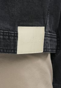 Sort denimjakke med et struktureret cremefarvet mærke, der har det prægede tekst "ONLY & SONS." Sømene er synlige langs kanterne.