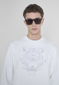Felpa bianca con una grafica di tigre grigia, caratterizzata da un collo a giro. Il modello indossa occhiali da sole scuri, con capelli corti e strutturati.