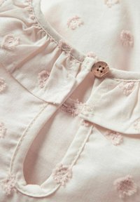 Camicia di un rosa chiaro con scollo arricciato, fiori ricamati a rilievo e un bottone di legno decorativo all'apertura frontale.