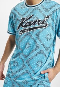 Hombre con una camisa de patrón de bandana azul con el logotipo "Kani est. 1989" en blanco y negro en el pecho.