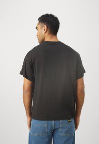 Sort bomulds t-shirt med korte ærmer, rund hals og lige kant. Har et lille logo nær den nederste kant.
