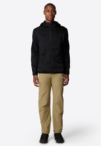 Chaqueta negra con capucha, con cierre frontal y tejido texturizado, combinada con pantalones cargo ligeros de color beige claro que tienen bolsillos con cremallera y costuras reforzadas.