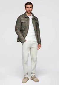 Uomo che indossa un giubbotto trapuntato oliva, un maglione a righe bianche, pantaloni beige chiaro e scarpe in suede beige, in piedi con una mano in tasca.