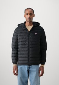 Juodas pūkinis švarkas su kapisonu, su quilted dizainu ir užtrauktuko uždarymu. Maža logo ant krūtinės, derinama su šviesiai mėlynomis džinsomis.