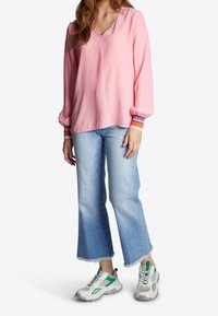 Blusa rosa a maniche lunghe con scollatura a V, caratterizzata da dettagli a righe sui polsini. Abbinata a jeans chiari con orlo sfilacciato e scarpe da ginnastica grigie con accenti verdi.