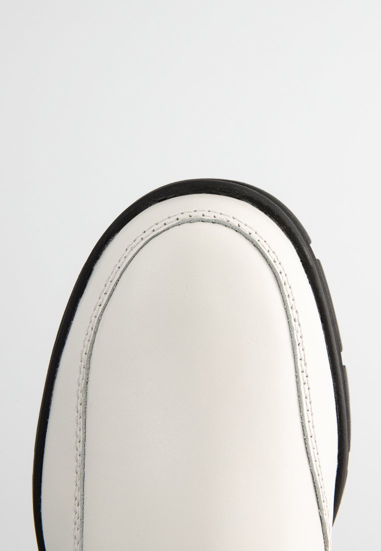 Weiße Lederschuhe mit schwarzer Gummisohle, runder Zehenform und sichtbaren Nähten am Rand. Glatte Textur, minimalistisches Design.