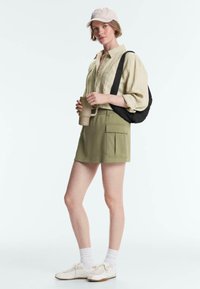 Femme portant une casquette beige, une chemise rayée, une jupe verte avec des poches, des chaussettes blanches et des baskets, tenant un mug de voyage beige et un sac à bandoulière noir.