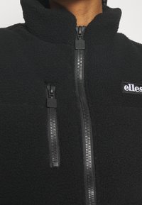Giacca in pile nera con due cerniere verticali nere e logo bianco "ellesse" sul petto, indossata con la cerniera completamente chiusa.