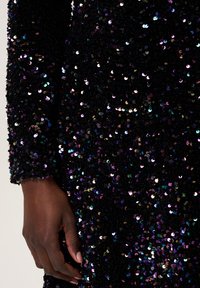 Tissu noir pailleté avec une surface texturée, présentant des sequins réfléchissants multicolores. Détail à manches longues avec placement de main visible.