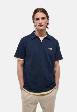 Jeune homme aux cheveux mi-longs portant un polo bleu marine à manches courtes par-dessus un t-shirt beige et un pantalon beige, les mains dans les poches.