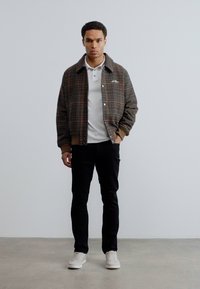 Καρό μπουφάν τύπου bomber σε καφέ και πράσινο, λευκό πόλο πουκάμισο, μαύρο παντελόνι και λευκά αθλητικά παπούτσια, στέκεται σε ανοιχτό γκρι πάτωμα μπροστά σε έναν απλό τοίχο.