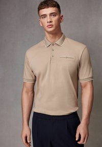 Beige polo-paita lyhyillä hihoilla ja kuvioidulla kauluksella, jossa on rintatasku ja erottuvat reunusompelut. Yhdistetty tummiin housuihin.